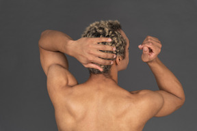 Vista posterior de un hombre afro sin camisa tocando su cabeza y levantando el brazo