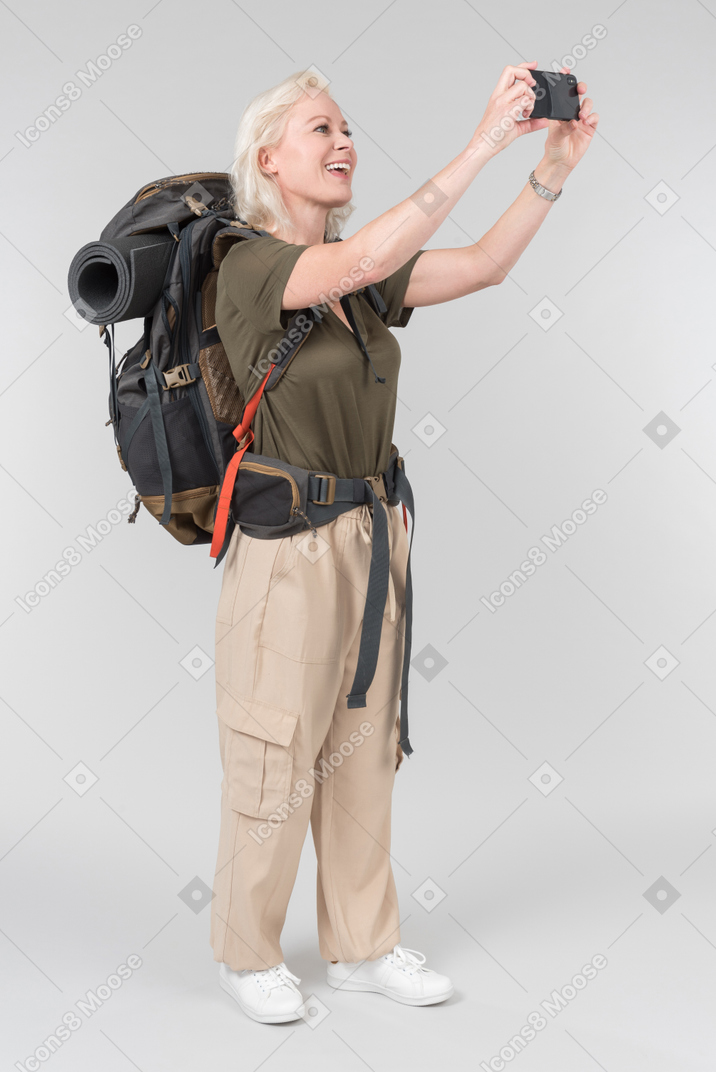 Mature female tourist carrying heavy backpack and making photos with smartphone