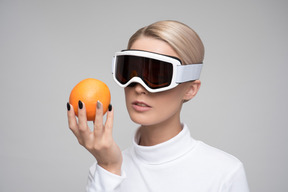 Jovem loira com óculos de esqui segurando uma laranja