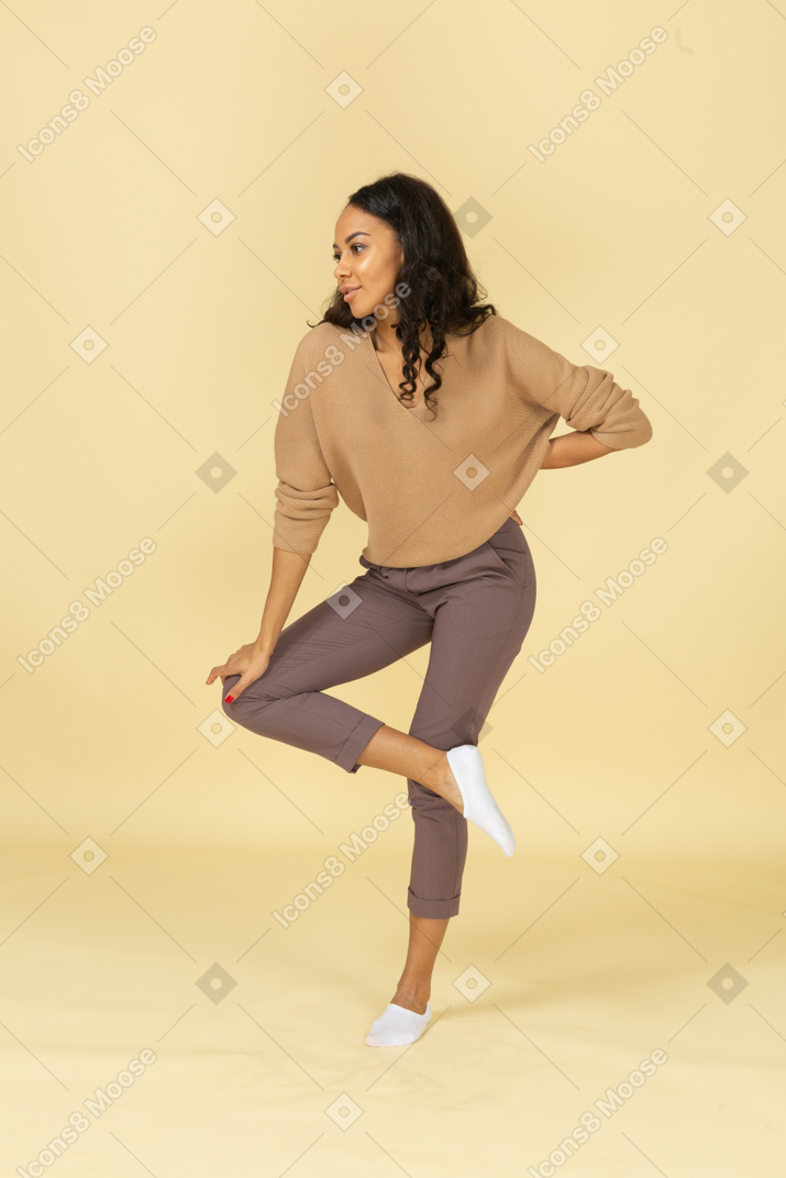 Vista de tres cuartos de una mujer joven de piel oscura que levanta la pierna mientras se inclina hacia adelante