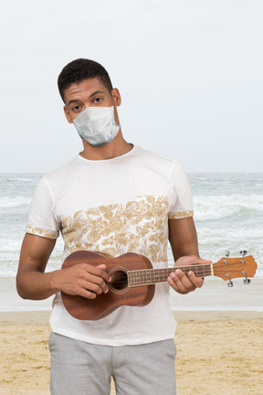 A man in a face mask playing a ukulele on the beach