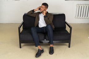 Front view of a perplexed young man sitting on a sofa and talking on his phone