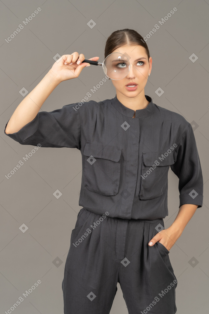 Front view of a puzzled young woman in a jumpsuit holding a magnifying glass