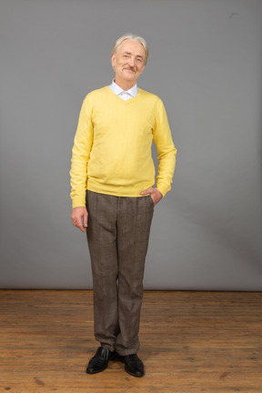 Vista frontal de un anciano complacido en un jersey amarillo poniendo la mano en el bolsillo y mirando a la cámara