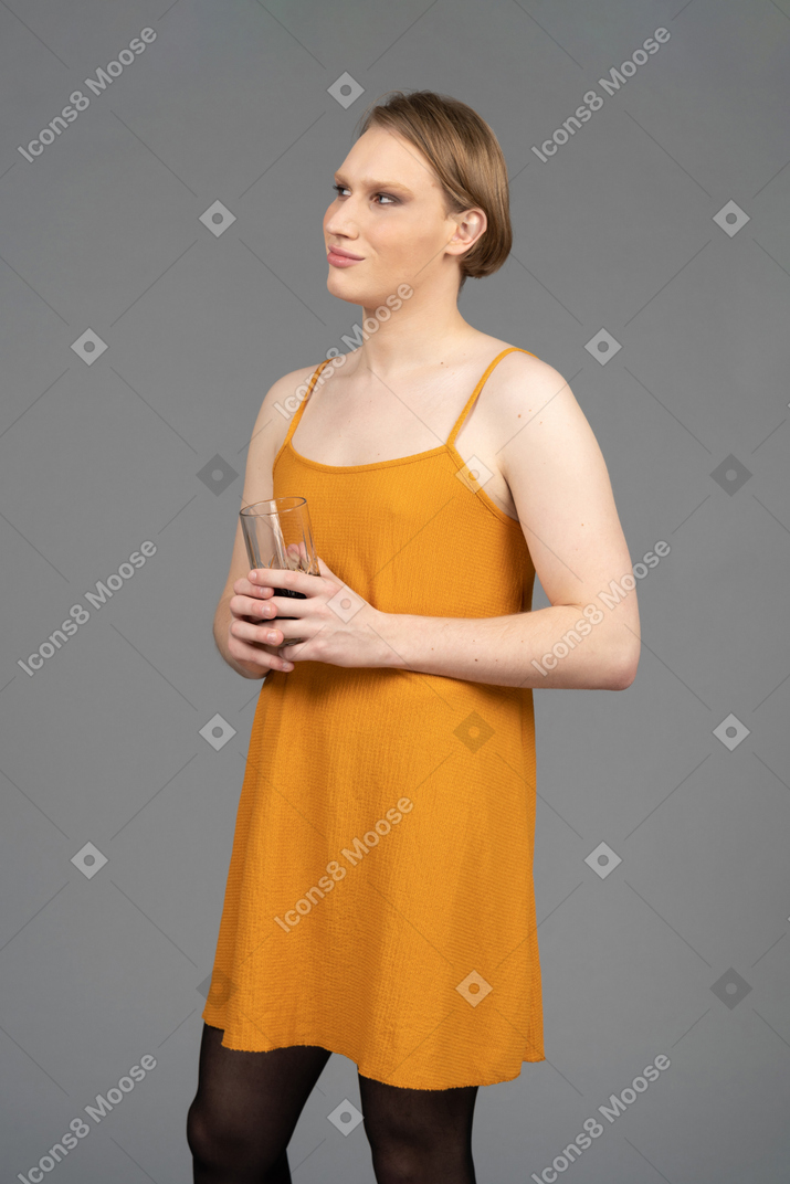 Молодой трансгендер в оранжевом платье держит стакан