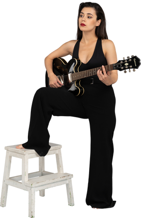 Вид в три четверти молодой женщины в черном костюме, держащей гитару и ставящей ногу на табурет