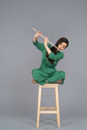 Toute la longueur d'une jeune femme jouant de la clarinette assise, les jambes croisées sur une chaise en bois