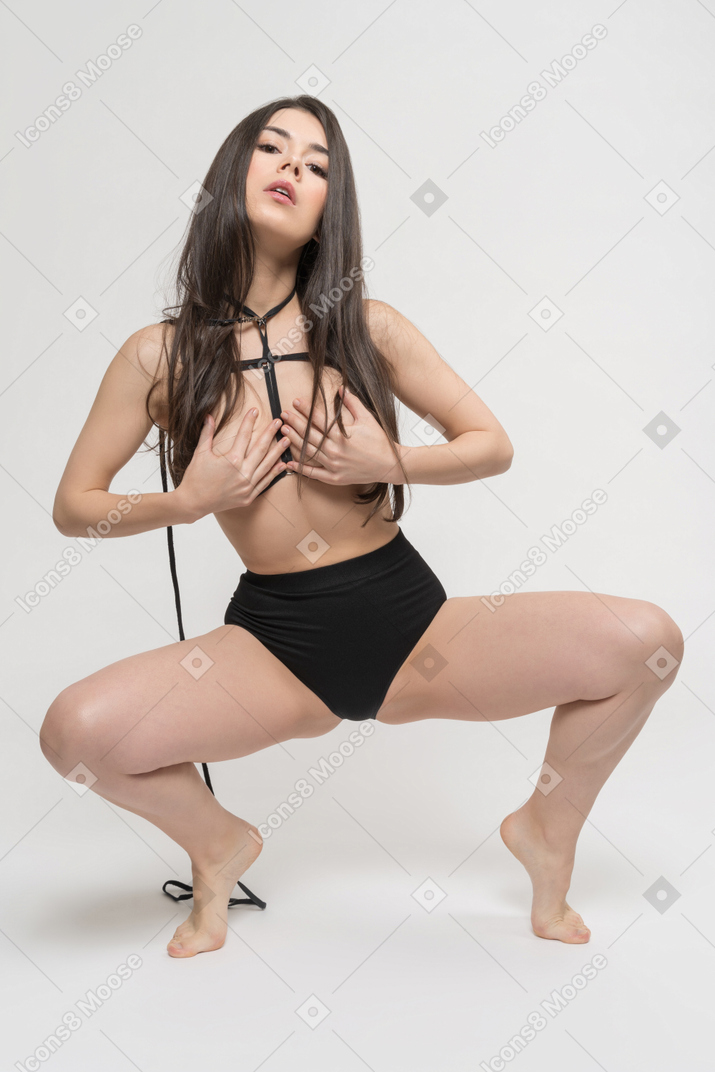 Front view of sexy young woman in harness sitting with legs spread and covering breast with hands