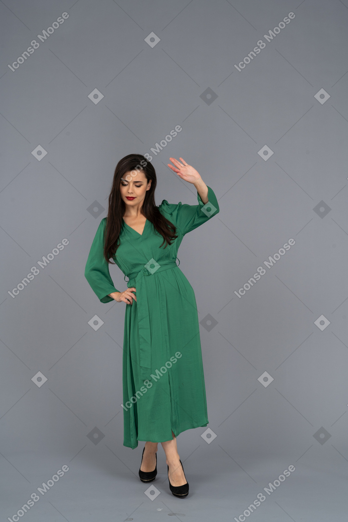 Vista frontal de una joven en vestido verde poniendo la mano en la cadera mientras levanta la mano