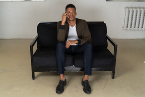 Front view of a perplexed young man sitting on a sofa and talking on his phone