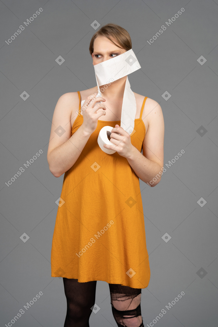 Young queer person in orange dress wrapping toilet paper around head