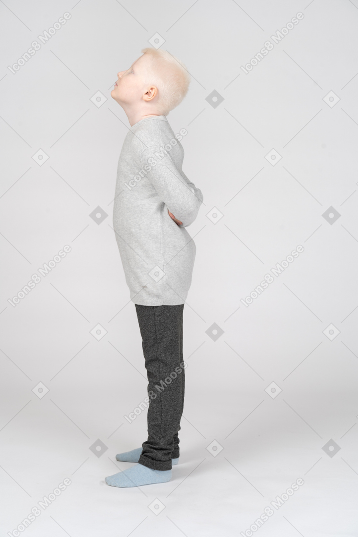 Un petit garçon blond dans des vêtements décontractés debout et se cachant les mains derrière