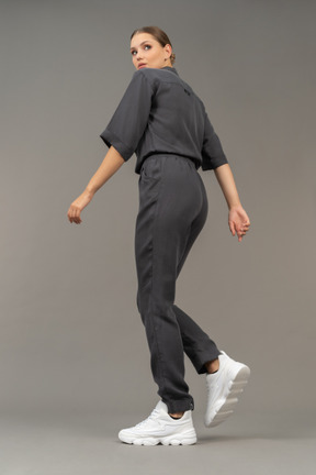 Three-quarter back view of a walking young woman in a jumpsuit looking aside