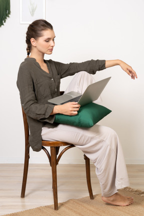 Vista lateral de uma jovem vestindo roupas de casa, sentada em uma cadeira com um laptop
