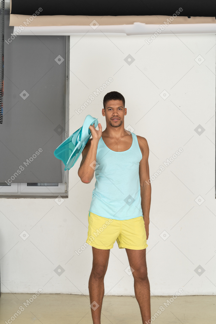 Front view of a man in yellow shorts and a blue tank top holding flippers