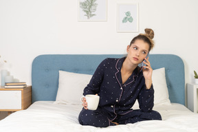 Frente a una mujer joven en pijama sentada en la cama sosteniendo la taza y teniendo una llamada telefónica