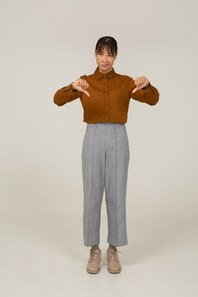Vista frontal de uma jovem mulher asiática de calça e blusa com os polegares para baixo