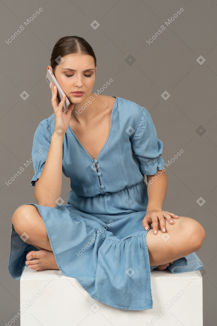Вид спереди молодой женщины, сидящей на кубе и разговаривающей по смартфону