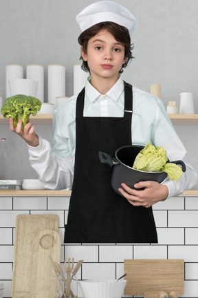 A boy in a chef's outfit holding broccoli