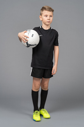 Vista de tres cuartos de un niño en uniforme de fútbol sosteniendo una pelota y mirando a un lado