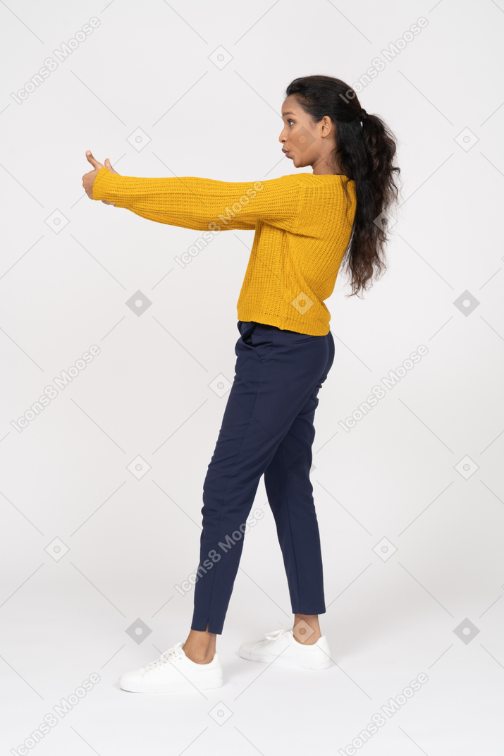 Vista lateral de una niña en ropa casual mostrando los pulgares hacia arriba y haciendo muecas