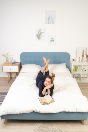 Вид спереди мечтающей молодой женщины, читающей книгу в постели