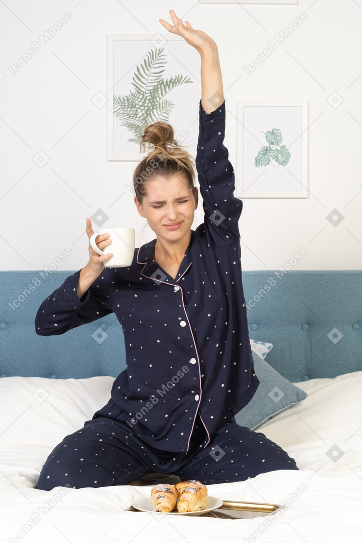 Front view of a stretching young lady in pajamas holding a cup of coffee while sitting in bed