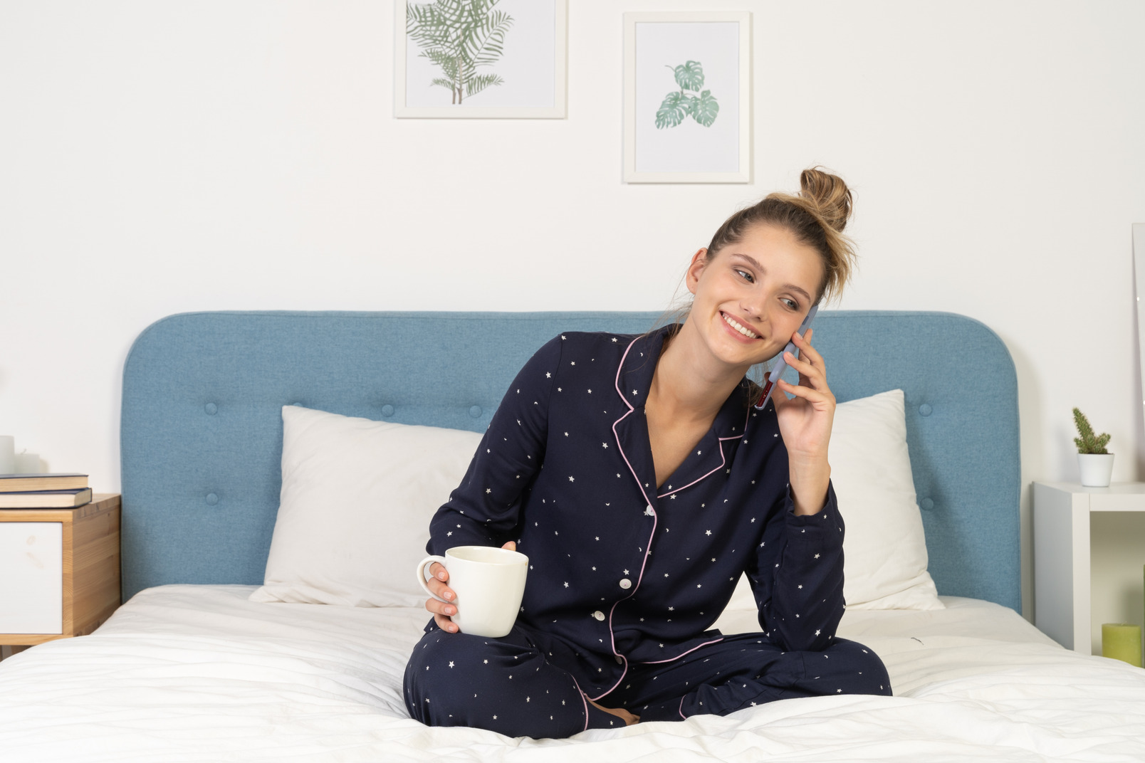 Front of a young female in pajama sitting in bed holding the cup and having a phone call