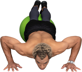 Vista frontal de un hombre afro sin camisa haciendo flexiones en una pelota de gimnasia