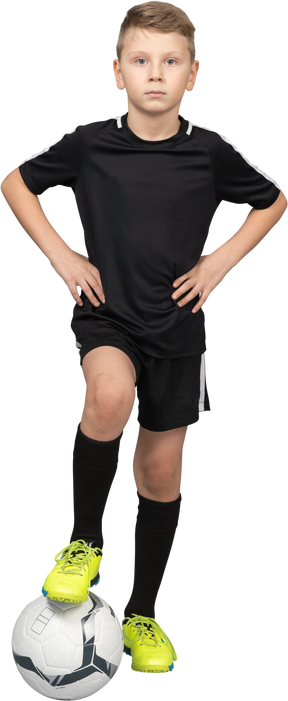 Vista frontal de un niño chico en uniforme de fútbol poniendo las manos en las caderas y el pie en la bola