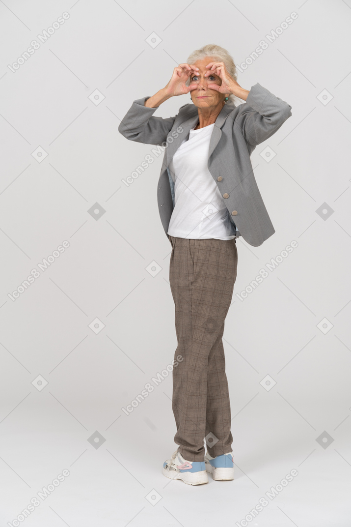 Vista frontal de una anciana en traje mirando a la cámara a través de binoculares imaginarios