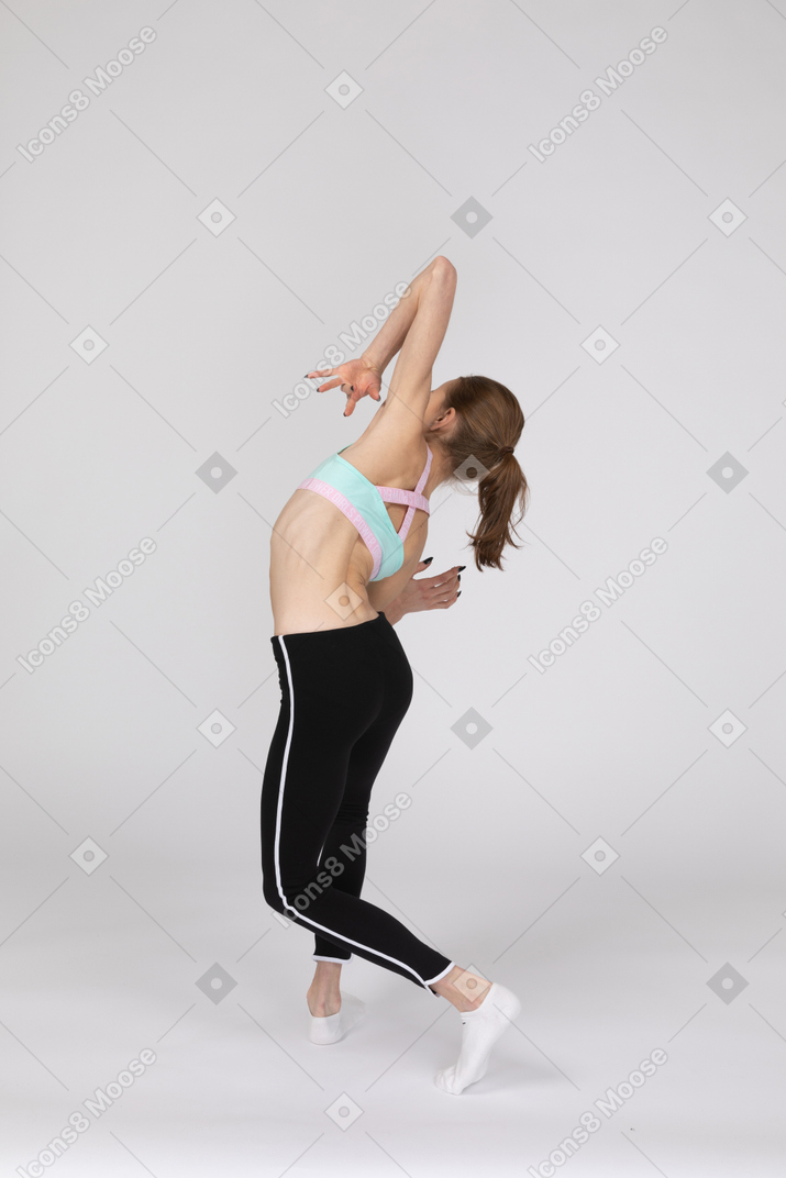 Vista posterior de tres cuartos de una elegante jovencita en ropa deportiva levantando las manos e inclinando