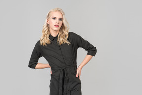 A young blond-haired person in a black dress  standing against the plain grey background