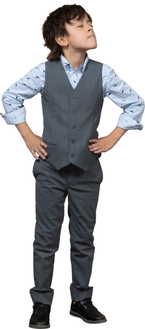 Front view of a cute boy in grey suit posing with hands on hips and looking up