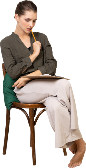 Вид спереди вдумчивой молодой женщины в домашней одежде, сидящей на стуле и делающей заметки