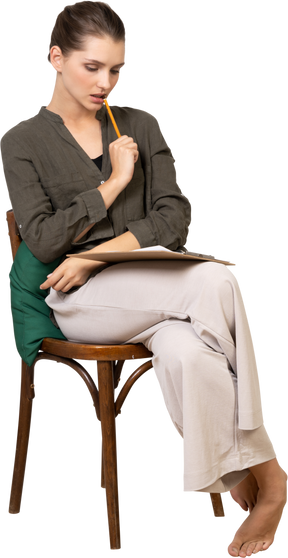 Front view of a thoughtful young woman wearing home clothes sitting on a chair and making notes