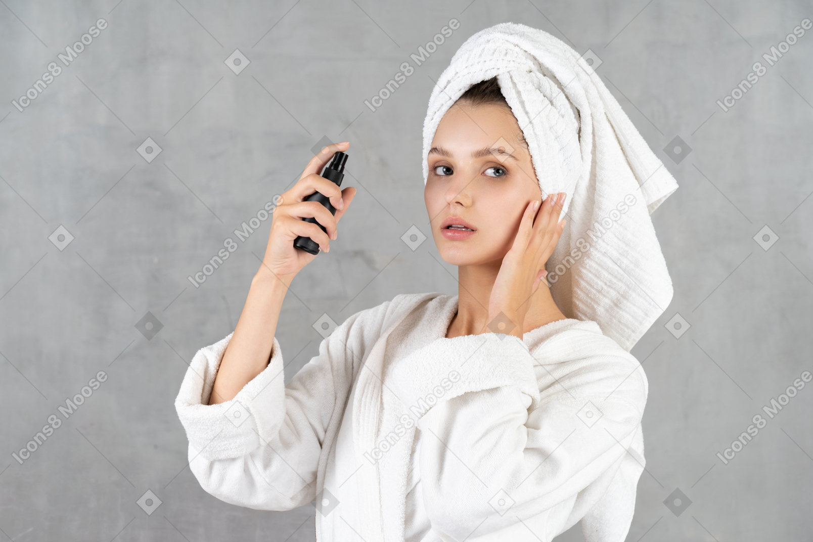 Woman in bathrobe spraying her face and touching her cheek
