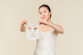 Giovane donna asiatica che osserva da vicino la maschera facciale che tiene con entrambe le mani