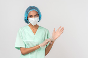 A young woman in a surgical hat and a face mask putting on gloves