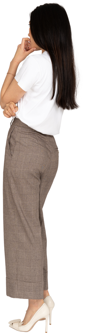 Vista posterior de tres cuartos de una joven pensativa en pantalones y camiseta tocando la barbilla