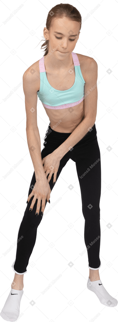 Vista frontal de una jovencita en ropa deportiva tocando la pierna e inclinándose hacia adelante