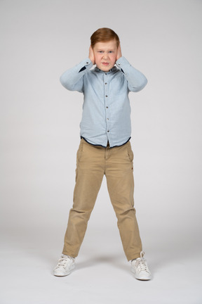 Vista frontal de un niño enojado cubriendo las orejas con las manos y mirando a la cámara