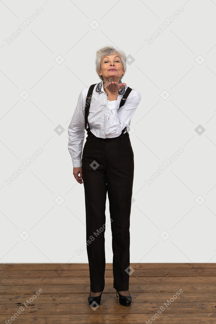 Front view of an old funny female in office clothes grimacing with her hands behind back sending air kiss