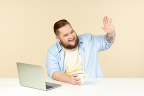 Cheerful young overweight man sitting at the table with laptop on it and welcoming someone