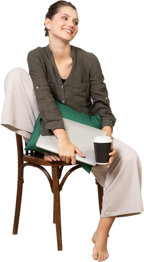 Front view of a smiling young woman sitting on a chair and holding her laptop & coffee cup