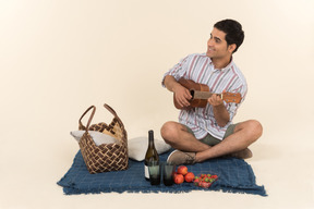 Молодой кавказский парень сидит на одеяле и играет на маленькой гитаре