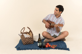 Young caucasian guy sitting on blanket and playing small guitar