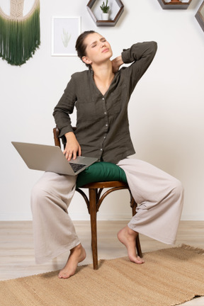 Front view of a tired young woman wearing home clothes sitting on a chair with a laptop