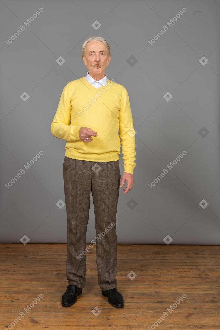 Вид спереди озадаченного старика в желтом пуловере, поднимающего руку и смотрящего в камеру