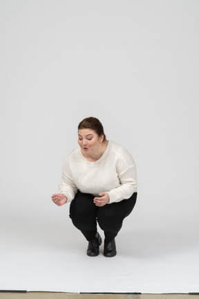 Женщина большого размера в белом свитере сидит на корточках, вид спереди
